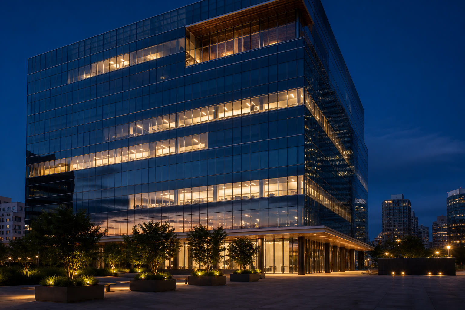 A modern commercial office tower at dusk, glass curtain wall lit warm from within against a deep blue twilight sky, seen across a landscaped plaza.